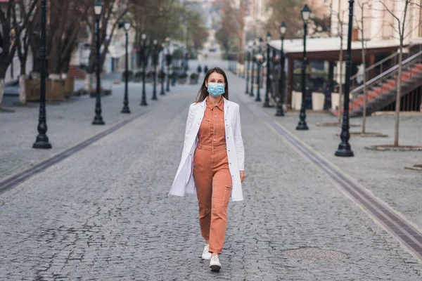 Turuncu elbiseli esmer bir kadın beyaz bir ceket giyiyor ve yüzünde koruyucu bir maske var.