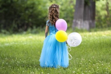 Görünümü tam uzunlukta portre sevimli küçük sarı uzun saçlı kız uzun mavi elbiseli renkli balonlar bulanık yeşil ağaçlar arka plan üzerinde çiçek açan alanda dönüyoruz. Mutlu çocukluk kavramı.