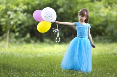 Küçük güzel uzun saçlı sarışın güzel uzun mavi gece elbisesi renkli balonlar bulanık yeşil ağaçlar arka plan üzerinde çiçek açan alanındaki parlak güneşli gün ayakta tutar. Mutlu çocukluk kavramı.