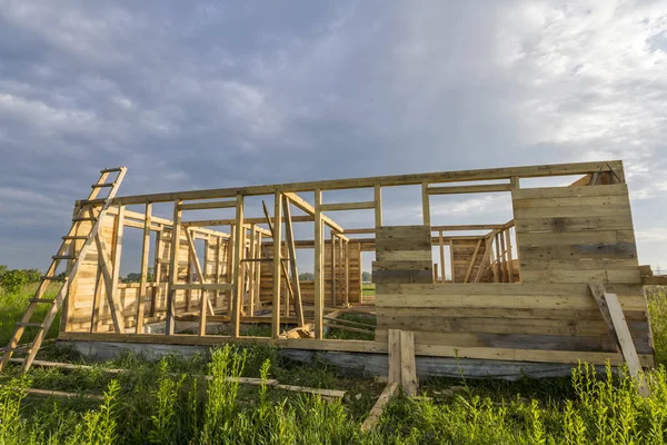 Unfinished ecological house under construction in green field on blue sky background. Trenches filled with cement and wooden walls with window openings of future comfortable cottage in quiet area.