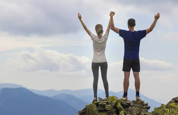 Back view of young couple, athletic boy and slim girl standing with raised arms on rocky mountain top enjoying breathtaking summer mountain view. Tourism, traveling and healthy lifestyle concept.