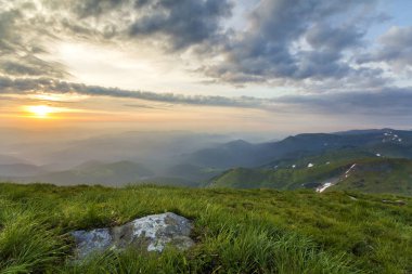 Geniş Yataklı güneş doğarken yaz. Parlak turuncu güneş mavi bulutlu gökyüzü büyük kaya ve uzak dağ ile yeşil çimenli tepeye üzerinde yükselterek sabah sis ile kaplı. Doğa kavramının güzellik.