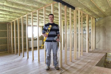 Genç yakışıklı alt düzey ve tornavida kamera büyük ışık Tavan odasında meşe zemin, yalıtılmış tavan ve ahşap çerçeve ile gelecekteki duvarlar için arıyorum. İnşaat ve tadilat kavramı.