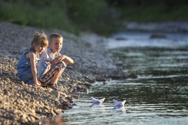 İki sarışın çocuk, erkek ve kız Nehri üzerinde beyaz kağıt tekne ile oynarken gülümseyen parlak yaz bulanık mavi arka plan üzerinde banka. Sevinçlerini ve mutlu çocukluk ve açık hava faaliyetleri kavramı ile oyun.