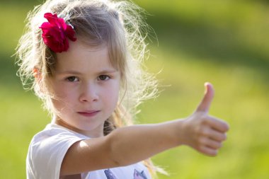 Küçük sarışın ciddi ile şirin kız beyaz tişört içinde saç başparmak-up jest fotoğraf bulanık parlak yeşil yaz arka plan üzerinde gösterilen Kırmızı gül portresi. Çocukluk kavramının güzellik.