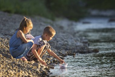 İki sevimli sarışın çocuk, erkek ve kız nehir kıyısında su beyaz kağıt tekne içinde parlak yaz gönderme mavi arka plan bulanık. Sevinçlerini ve mutlu çocukluk ve açık hava faaliyetleri kavramı ile oyun.