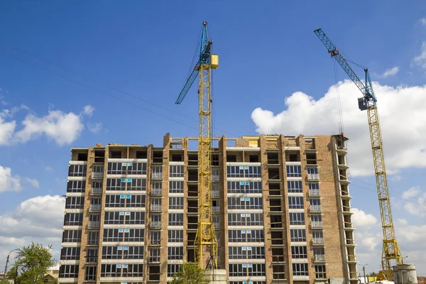 Building site with two industrial tower cranes working at construction of new brick tall building on bright blue sky and green trees background.