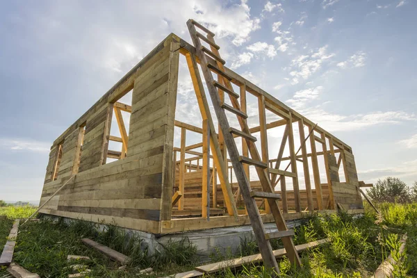 Unfinished ecological house under construction in green field on blue sky background. Trenches filled with cement and wooden walls with window openings of future comfortable cottage in quiet area.