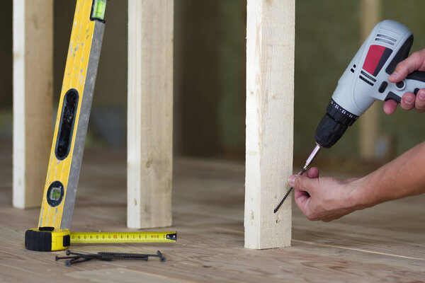 Close-up of worker's hands with screwdriver on background of professional tools and wooden frame for future wall in unfinished attic room under reconstruction. Renovation and improvement concept.