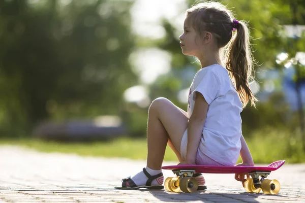 Oldukça küçük uzun saçlı sarışın kız beyaz giyim kaykay üzerinde bulanık bokeh arka plan üzerinde parlak yaz gününde oturma portre profil. Çocuk Aktiviteleri ve aktif yaşam tarzı kavramı.