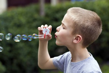 Profil sevimli yakışıklı küçük sarışın çocuk çocuk komik ciddi ifade üzerinde açık havada renkli şeffaf sabun köpüğü üfleme ile yeşil yaz arka plan bulanık. Sevinç dikkatsiz çocukluk kavramının.