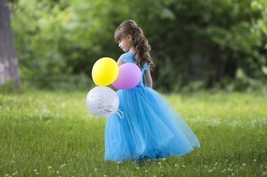 Renkli balonları bulanık yeşil ağaçlar arka plan üzerinde çiçek açan alanda tam uzunlukta portre sevimli küçük sarı uzun saçlı kız uzun mavi elbiseli profil. Mutlu çocukluk kavramı.