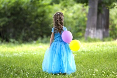 Görünümü tam uzunlukta portre sevimli küçük sarı uzun saçlı kız uzun mavi elbiseli renkli balonlar bulanık yeşil ağaçlar arka plan üzerinde çiçek açan alanda dönüyoruz. Mutlu çocukluk kavramı.