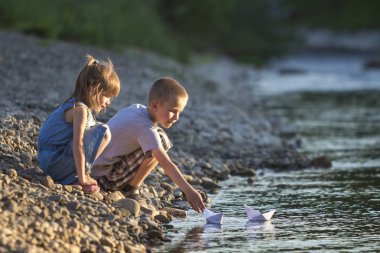 İki sevimli sarışın çocuk, erkek ve kız nehir kıyısında su beyaz kağıt tekne içinde parlak yaz gönderme mavi arka plan bulanık. Sevinçlerini ve mutlu çocukluk ve açık hava faaliyetleri kavramı ile oyun.