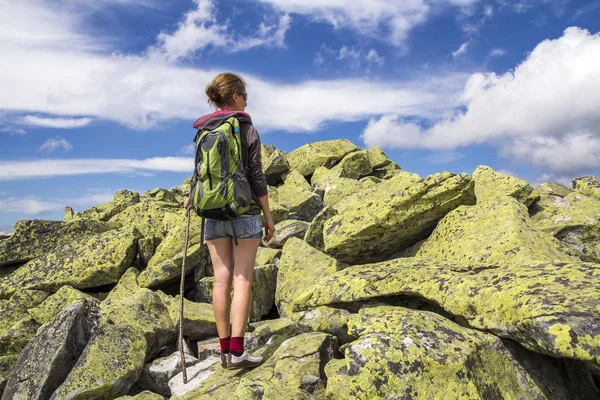 Slim athletic blond tourist hiker girl with stick and backpack climbing lit by sun high rocky mountain on bright blue sky background. Tourism, traveling, hiking and healthy lifestyle concept.