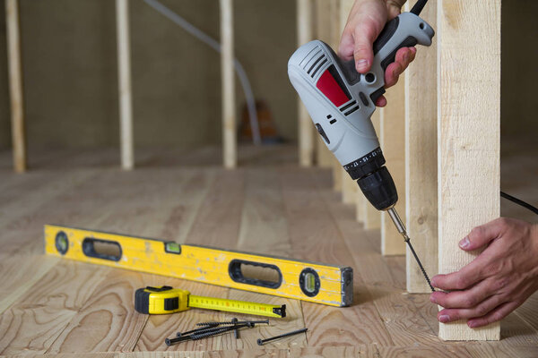 Close-up of worker's hands with screwdriver on background of professional tools and wooden frame for future wall in unfinished attic room under reconstruction. Renovation and improvement concept.