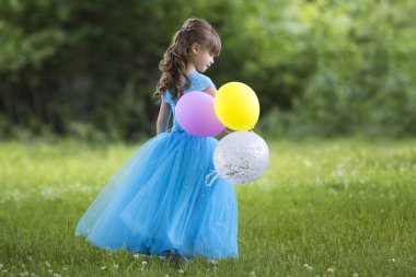 Renkli balonları bulanık yeşil ağaçlar arka plan üzerinde çiçek açan alanda tam uzunlukta portre sevimli küçük sarı uzun saçlı kız uzun mavi elbiseli profil. Mutlu çocukluk kavramı.