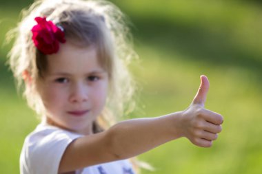 Sevimli küçük sarışın ciddi kız beyaz T-shirt saç başparmak-up jest fotoğraf bulanık parlak yeşil yaz arka plan üzerinde gösterilen Kırmızı gül ile bulanık portresi. Çocukluk kavramının güzellik.