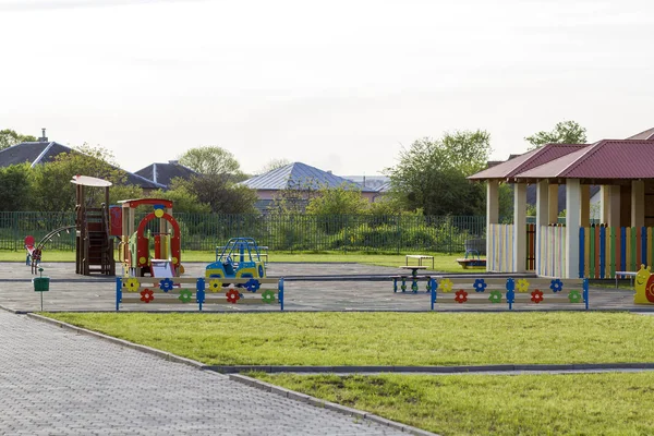 Beautiful playground in kindergarten with bright new alcove with red tile roof and multicolored low fence, green lawn, swings, slides and sandbox. Perfect place for children activities outdoors.