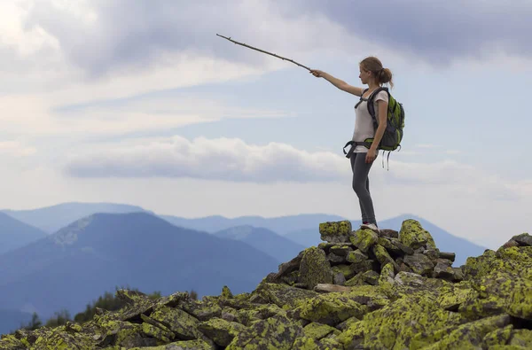 Young slim blond tourist girl with backpack points with stick at foggy mountain range panorama standing on rocky top on bright blue morning sky background. Tourism, traveling and climbing concept.