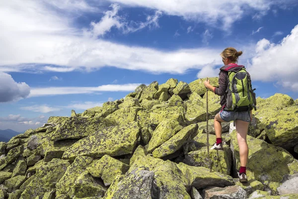 Slim athletic blond tourist hiker girl with stick and backpack climbing lit by sun high rocky mountain on bright blue sky background. Tourism, traveling, hiking and healthy lifestyle concept.