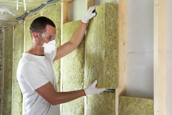 Worker in protective goggles and respirator insulating rock wool insulation in wooden frame for future house walls for cold barrier. Comfortable warm home, economy, construction and renovation concept