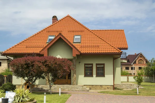 New nice one-story cottage with shingle roof, balcony, plastic attic windows, paved yard, green lawn and decorative blooming trees on bright blue sky background. Real estate property concept.