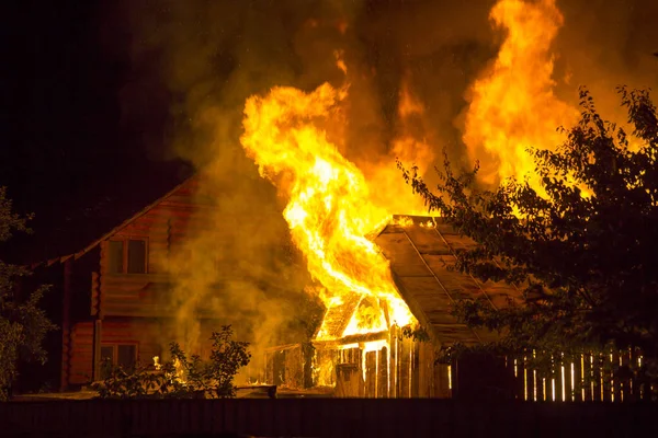 木造住宅を燃焼します 明るいオレンジ色の炎と暗い空 木のシルエットと住宅隣人コテージ背景に瓦屋根の下から濃い煙 災害や危険の概念 — ストック写真