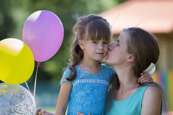 Playful beautiful blond small girl with colorful balloons in nice blue dress kissing her young mother on blurred summer bokeh background. Perfect family relations, happiness, love and care concept.