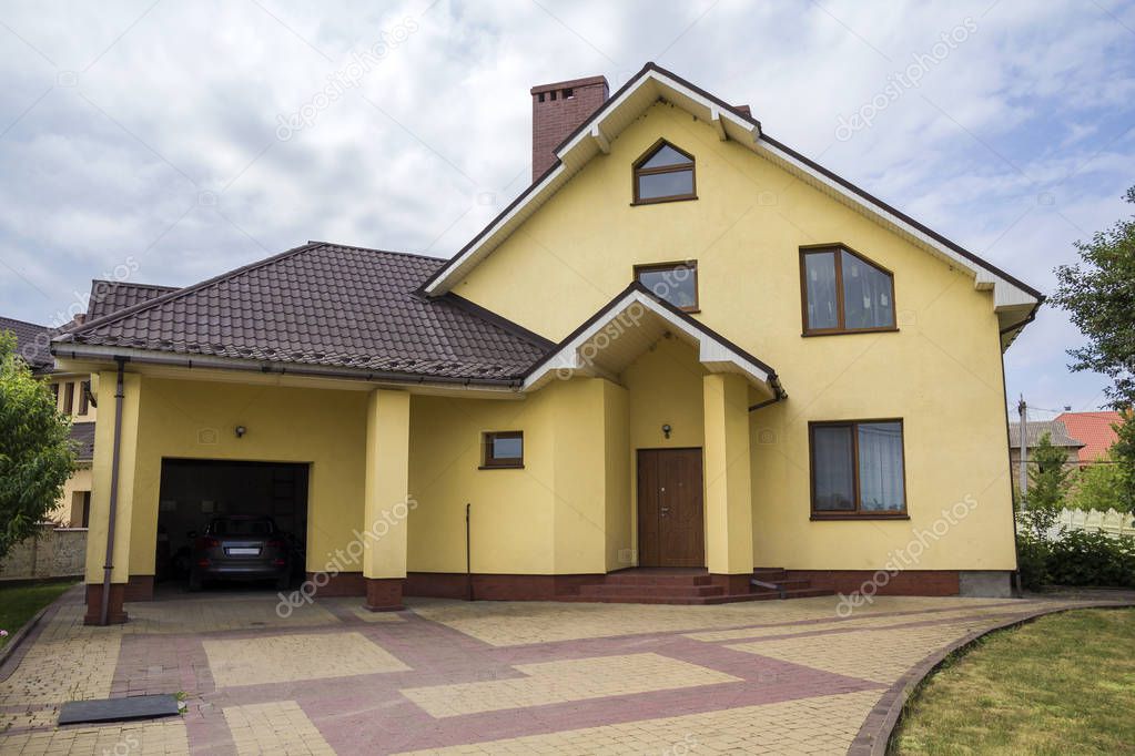 New nice yellow two-story residential cottage with shingle roof, plastic windows, high chimney, paved yard, car in attached garage on blue sky background. Real estate property and prosperity concept.