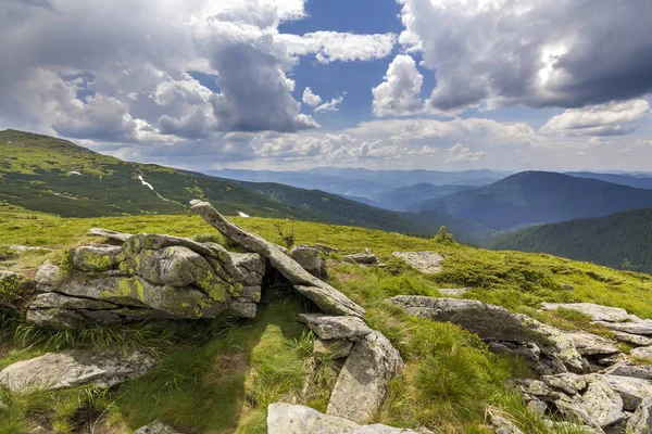 Wide panorama of lit by sun mountain plateau with green grass, patches of snow and big boulders on distant mountains under cloudy sky background. Beauty of nature, tourism and traveling concept.