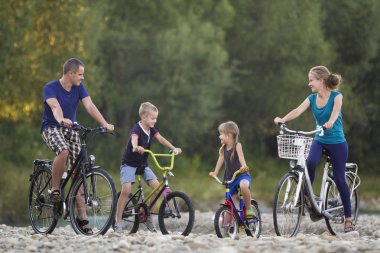 Genç mutlu anne, baba ve iki sevimli sarışın çocuk, çocuk ve kız çakıllı nehir kıyısında bulanık parlak yaz gün arka plan üzerinde bisiklet sürme. Aktif yaşam tarzı ve aile eğlence kavramı.