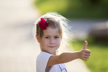 Küçük sarışın ciddi ile şirin kız beyaz tişört içinde saç başparmak-up jest fotoğraf bulanık parlak yeşil yaz arka plan üzerinde gösterilen Kırmızı gül portresi. Çocukluk kavramının güzellik.