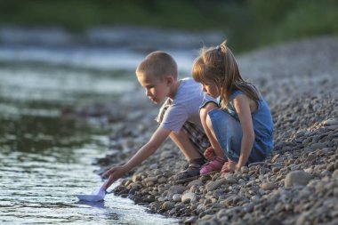 İki sevimli sarışın çocuk, erkek ve kız nehir kıyısında su beyaz kağıt tekne içinde parlak yaz gönderme mavi arka plan bulanık. Sevinçlerini ve mutlu çocukluk ve açık hava faaliyetleri kavramı ile oyun.
