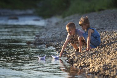 İki sevimli sarışın çocuk, erkek ve kız nehir kıyısında su beyaz kağıt tekne içinde parlak yaz gönderme mavi arka plan bulanık. Sevinçlerini ve mutlu çocukluk ve açık hava faaliyetleri kavramı ile oyun.
