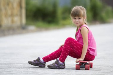 Genç ve güzel uzun saçlı sarışın çocuk kız bulanık güneşli yaz yeşil zemin üzerine döşeli banliyö sokakta kaykay üzerinde oturan rahat pembe kılığına girmiş. Çocuk Aktiviteleri, Oyunlar ve eğlence kavramı.