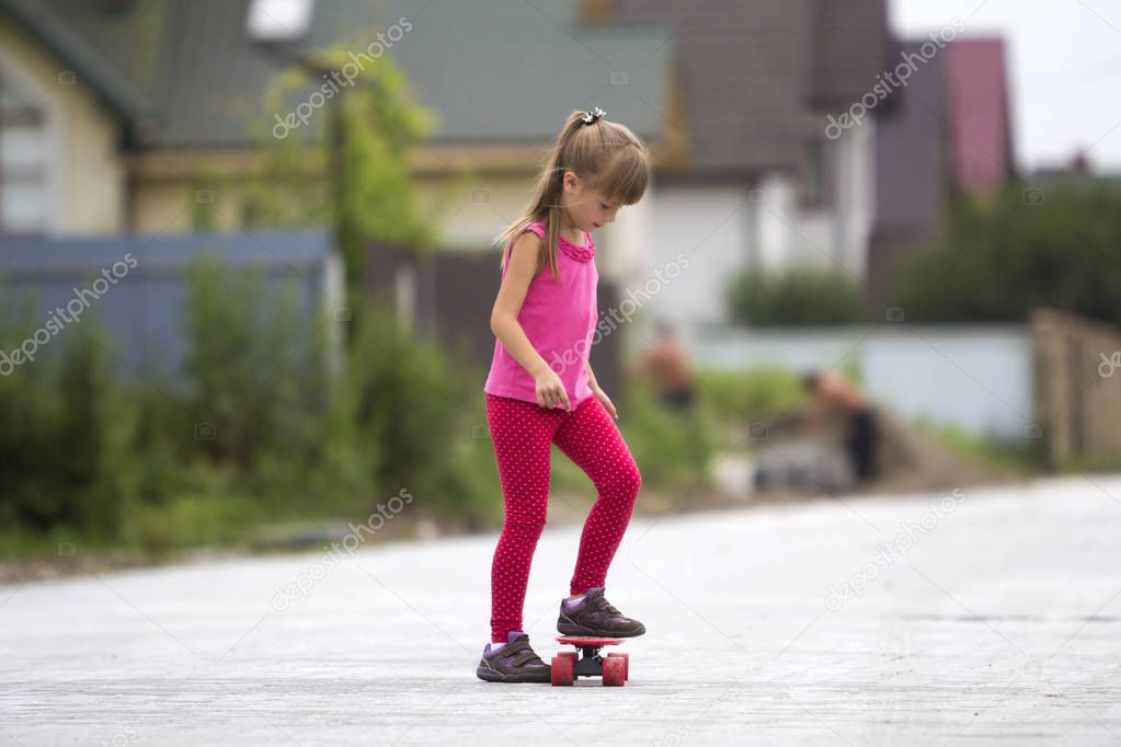 Pretty young long-haired blond child girl in casual clothing stand smiling on skateboard on lit by summer sun bright suburb street on blurred background. Children activities, games and fun concept.