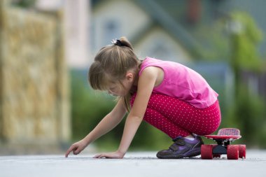 Genç ve güzel uzun saçlı sarışın çocuk kız bulanık güneşli yaz yeşil zemin üzerine döşeli banliyö sokakta kaykay üzerinde oturan rahat pembe kılığına girmiş. Çocuk Aktiviteleri, Oyunlar ve eğlence kavramı.