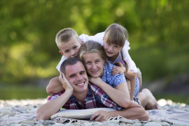 Genç mutlu anne, baba ve iki sevimli sarışın çocuk, erkek ve kız yeşil bokeh arka plan ile parlak yaz gününde aile portresi. Mutlu aile ilişkileri, sevgi, bakım ve mükemmel bir tatil kavramı