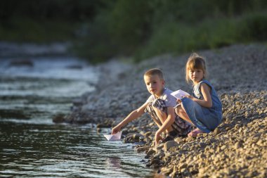 İki sevimli sarışın çocuk, erkek ve kız nehir kıyısında su beyaz kağıt tekne içinde parlak yaz gönderme mavi arka plan bulanık. Sevinçlerini ve mutlu çocukluk ve açık hava faaliyetleri kavramı ile oyun.