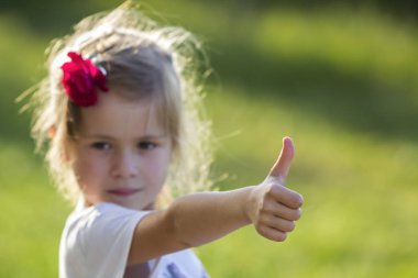 Gri gözleri ve kırmızı gül kamera için başparmak-up jest parlak bulanık yeşil açık havada arka plan üzerinde gösterilen güzel uzun saçlı küçük sevimli sarışın kız portresi. Mutlu çocukluk kavramı.