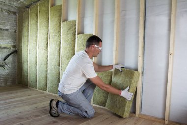 Worker in protective goggles and respirator insulating rock wool insulation in wooden frame for future house walls for cold barrier. Comfortable warm home, economy, construction and renovation concept clipart