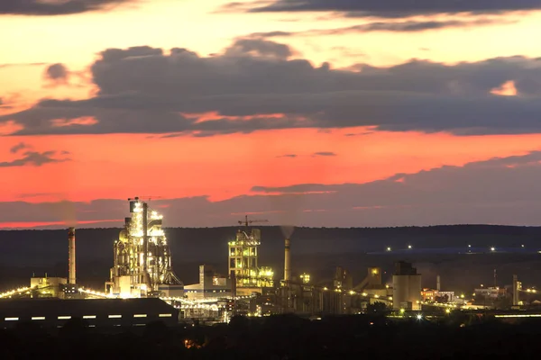 Panorama night view of brightly lit modern cement plant and power station in Ivano-Frankivsk, Ukraine under dramatic red sky on distant dark green hills background. Technology and ecological problems.