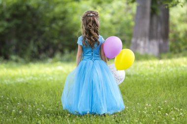 Görünümü tam uzunlukta portre sevimli küçük sarı uzun saçlı kız uzun mavi elbiseli renkli balonlar bulanık yeşil ağaçlar arka plan üzerinde çiçek açan alanda dönüyoruz. Mutlu çocukluk kavramı.
