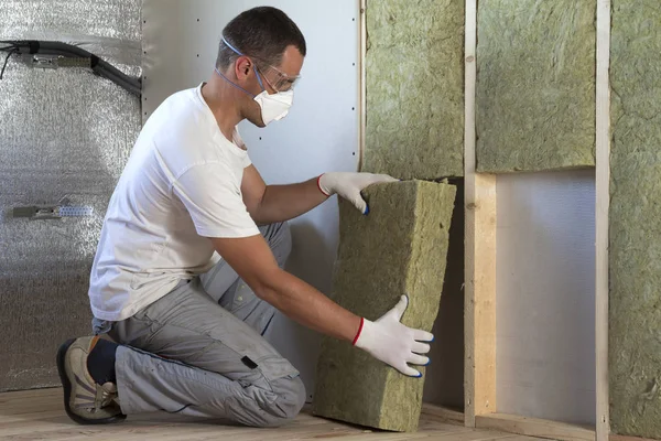 Worker in protective goggles and respirator insulating rock wool insulation in wooden frame for future house walls for cold barrier. Comfortable warm home, economy, construction and renovation concept