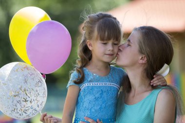 Oynak güzel sarışın küçük kız güzel mavi elbiseli Genç annesi öpüşme bulanık yaz bokeh arka plan üzerinde renkli balonlar ile. Mükemmel aile ilişkileri, mutluluk, sevgi ve bakım kavramı.