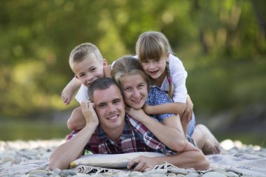 Genç mutlu anne, baba ve iki sevimli sarışın çocuk, erkek ve kız yeşil bokeh arka plan ile parlak yaz gününde aile portresi. Mutlu aile ilişkileri, sevgi, bakım ve mükemmel bir tatil kavramı