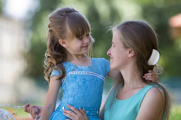 Playful beautiful small girl with long blond hair in nice blue dress hugging her mum\'s neck and looking at her with smile on blurred bokeh background. Perfect family relations, love and care concept.