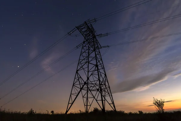 Transmission and long distance distribution of electricity concept. Angled view of high voltage tower with electric power lines stretching on dark blue starry sky background.