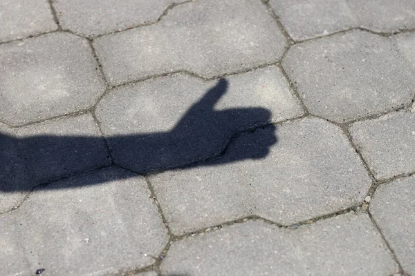 Close-up shot of shadow silhouette of human arm and hand with thumb-up success gesture on gray pavement lit by bright sun.